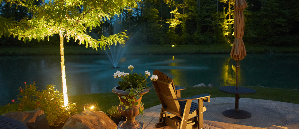 back yard patio with pond with chairs and trees that are illuminated at night time 