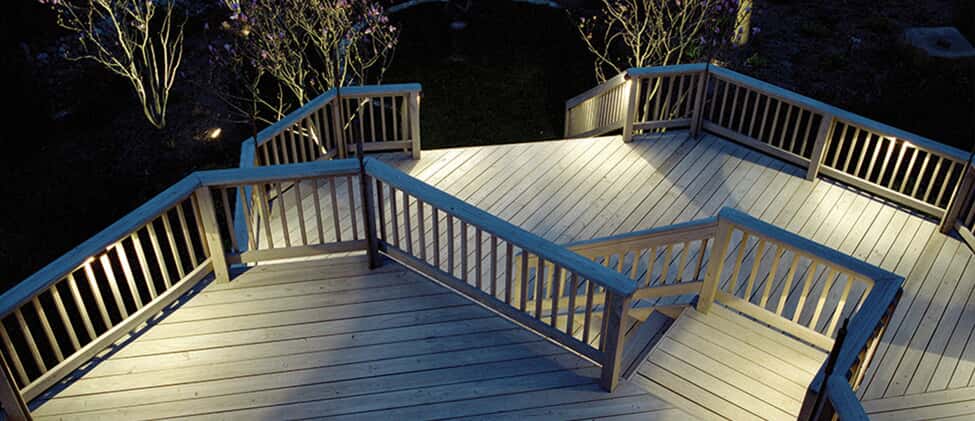 multi-level wood deck with stairs that has illumination lighting at night time leading down to back yard