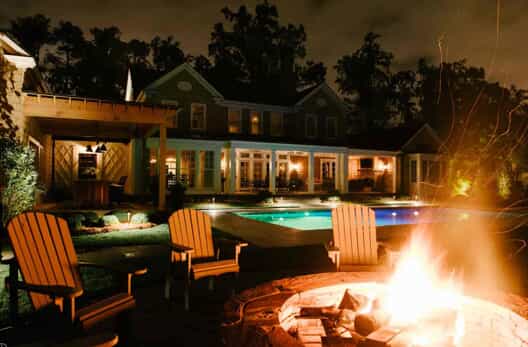 firepit, pool, landscaping, and home lit by outdoor lighting